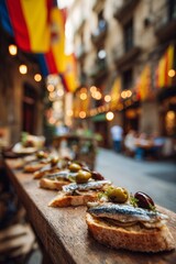 Fototapeta premium Spanish pintxos with anchovy, green olive, and red pepper on bread, served on narrow wooden bar counter, open to a stone alleyway with hanging flags and rustic village charm in background.