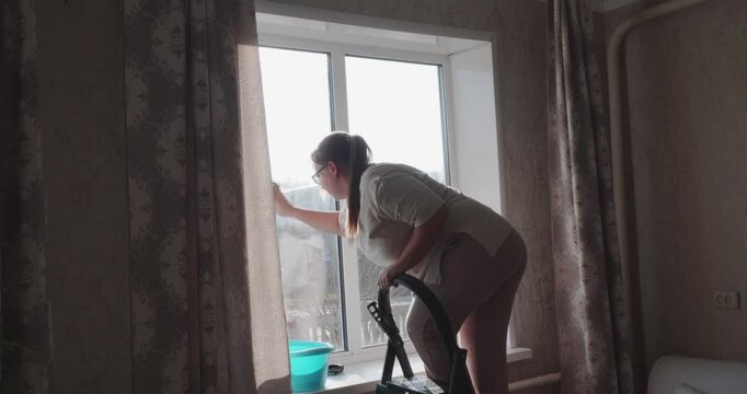 A hardworking housewife washes a large window with a rag and soapy water in a cozy room. The woman diligently performs her daily chores, maintaining the house with care and neatness.