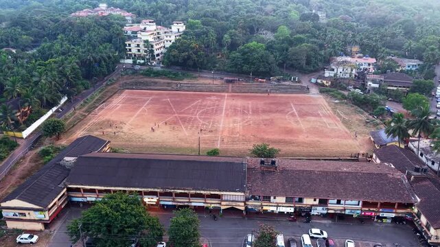 Aerial overhead view of large dusty football field with children playing soccer, marked pitch lines visible on red soil ground, community sports activity in open playground, energetic youth movement, 