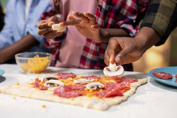 Close up of black young family making homemade pizza together and sprinkling mozzarella and pepperoni. Cozy weekend activity emphasizes bonding through delicious food, sharing laughter.