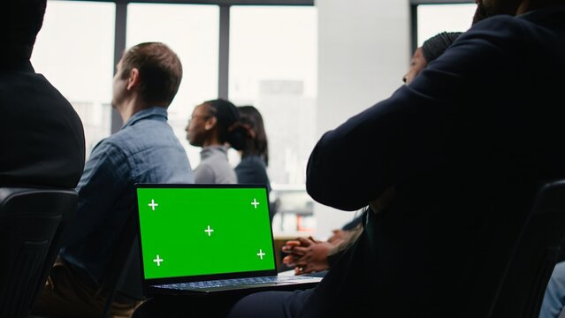 Green screen laptop during business event in conference hall with chairs filled by professionals. Guest speaker on stage leads panel discussion, encouraging audience engagement.
