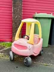 Abandoned plastic toy car left on pavement near garbage bins and garage doors, discarded children&rsquo;s ride-on toy, urban waste and neglected objects concept.