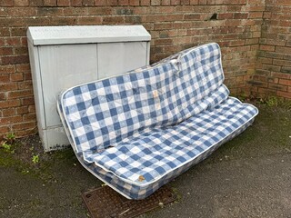 Discarded blue and white checkered mattress placed outdoors against a brick wall, abandoned bedding on pavement, urban waste and illegal dumping concept.