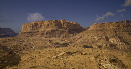 Expansive desert terrain showcases dramatic rock formations and cliffs bathed in sunlight. The atmosphere is tranquil with a few clouds dotting the bright blue sky, highlighting the vastness.