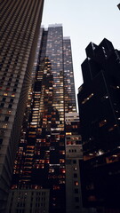 Skyscraper windows glowing at dusk, mirrored facades and warm office lights define corporate mood and dense downtown skyline, vertical perspective emphasizes