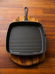 A black cast iron pan sits on a wooden cutting board. The pan is empty and the cutting board is made of wood. Solid metal tool for frying with ridges. Heavy duty kitchen utensil.