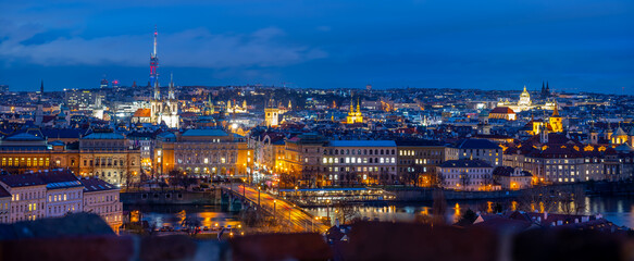 People enjoy the evening view of Prague. The city lights are bright against the dark sky. The skyline shows buildings, rivers, and bridges with reflections on the water.