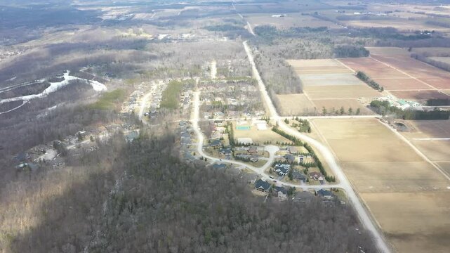 High altitude drone footage slowly flying over a suburban community nestled between a bare winter forest with patches of snow and expansive agricultural fields in the countryside
