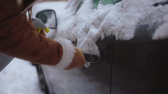 A close-up shot of a person using a de-icer spray and a key to unlock a car door covered in a thick layer of snow and ice during winter.
