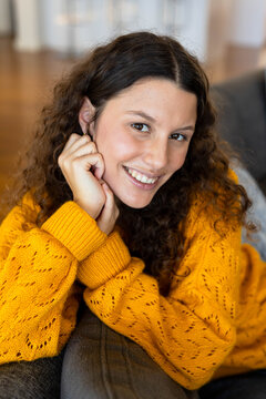Woman reclining on gray couch in living room, wearing mustard knit, hoop earring, wooden floor