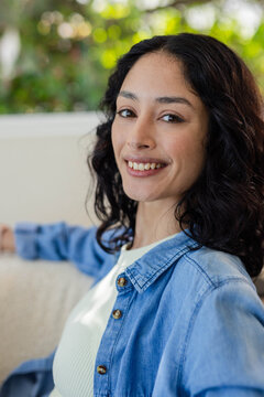 Female sitting on sofa in living room facing camera, wearing denim shirt and white ribbed top