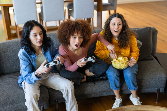 Diverse female friends playing game on sofa at home holding white controllers, sharing glass bowl