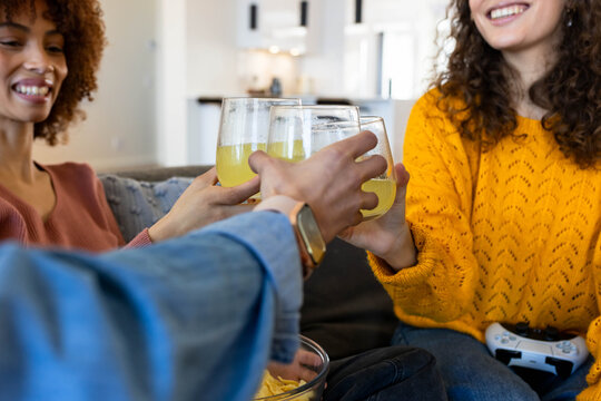 Diverse female friends clinking stemless glasses while sharing chips and controller on couch