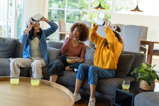 Diverse female friends sitting on gray sofa at home, trying VR headsets while snacking from bowl