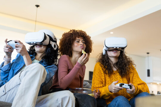 Diverse female friends wearing VR headsets and holding controllers while snacking on chips on sofa