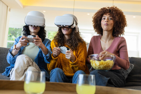 Three female friends gaming with VR headsets and controllers on sofa holding bowl of chips