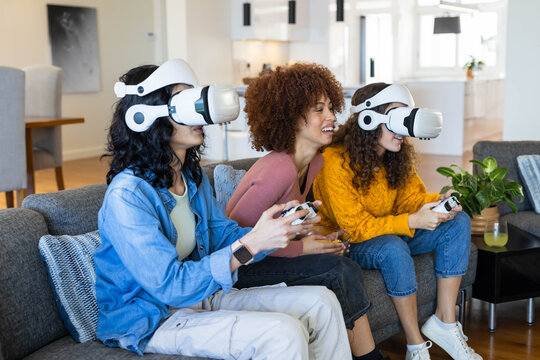 Diverse female friends sitting on gray sofa, two wearing white VR headsets holding controllers