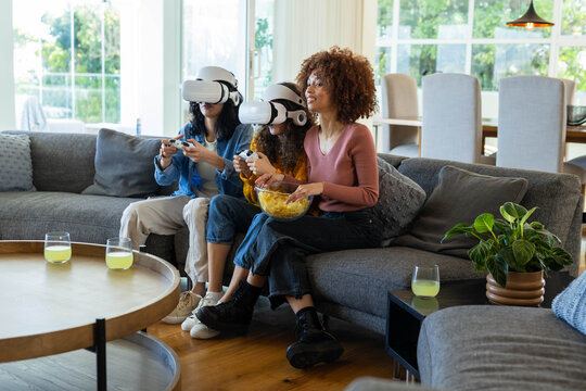 Diverse female friends sitting on sofa in living room with VR headsets holding remotes and popcorn