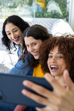 Female friends leaning together on patterned couch near window, taking selfie with smartphone
