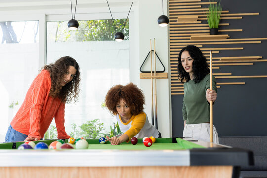 Diverse female friends playing on pool table, lining up shot in modern game room with cues