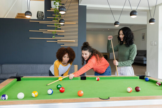 Diverse female friends lining up shot over green-felt pool table in modern living room with cues