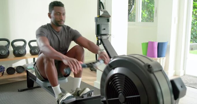 Securing foot straps African American man in gray tee powering rower in home gym boosting fitness