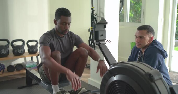 African American man rowing checking watch, sipping water while friend coaching at gym, copy space