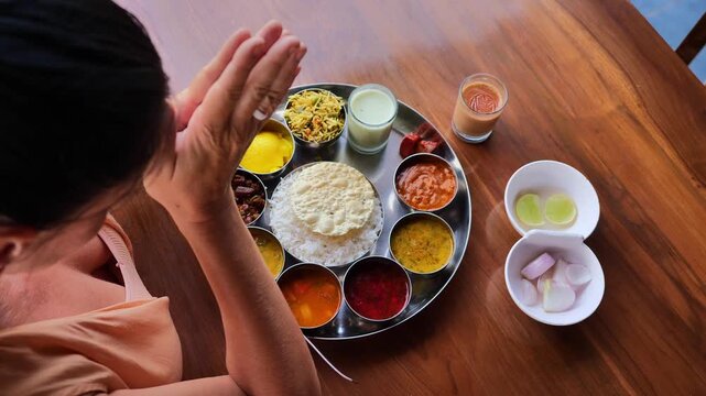 Overhead view of a woman in peach dress sitting at a wooden table with hands raised in excitement before eating Indian thali meal, showcasing cultural appreciation, joy of traditional Indian food, and