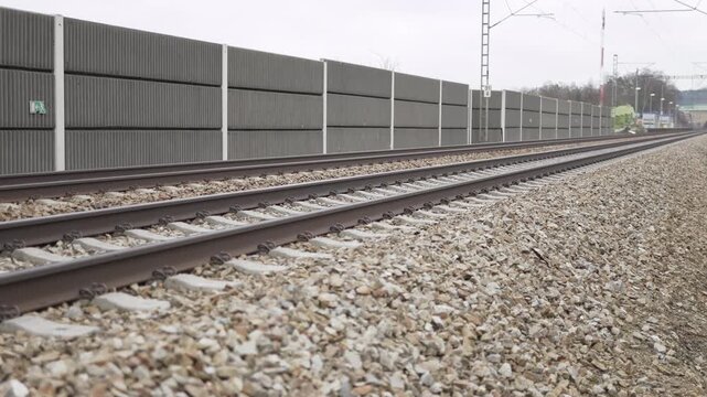 Railway track view.  Gravel ballast visible.  Noise barrier present.  Linear perspective.  Transportation infrastructure.  Industrial scene.  Motion blur effect.  Focus on rails.  Background details