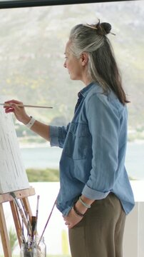 Vertical video: Sunlight bathing senior painter at easel using thin brush, capturing view, denim