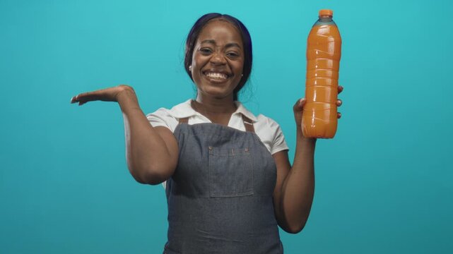 Woman smiling and holding an orange juice bottle with palm up presenting, wearing denim apron in studio; refreshment health joy.