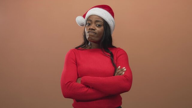 Woman wearing santa hat folds arms in studio with brown backdrop and red sweater visible; holiday annoyance.
