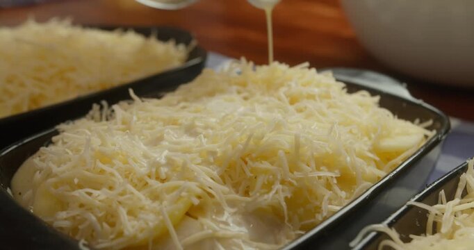 Chef pours creamy white b chamel sauce over layered sliced potatoes and cheese in cast iron baking dishes. Close-up of cooking process, homemade comfort food, classic French sauce,