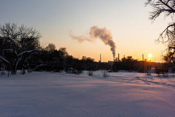 Stunning winter snowfield meets industrial sunset haze with dark silhouettes of bare trees along a frozen riverbank