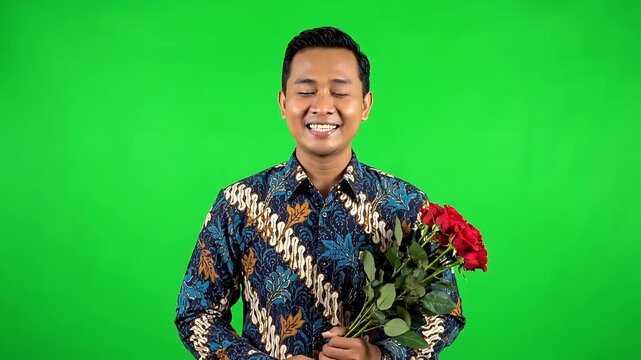 Joyful man in batik shirt holding red roses, eyes closed, on green screen background