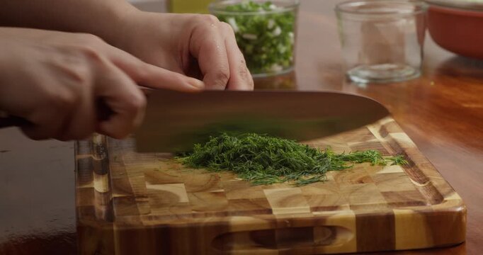 Close-up of fresh herbs being finely chopped on a wooden cutting board on a rustic wooden table. Chef s hands preparing spring onions, parsley, and dill for homemade cooking in natural light.