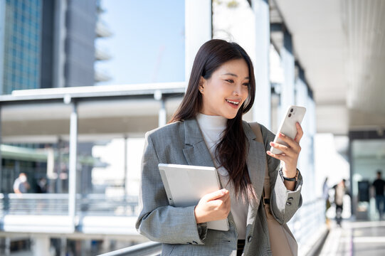 Pretty asian woman student office worker holding tablet looking at phone and standing on footbridge.