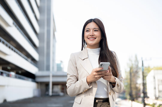 Pretty asian woman in beige blazer holding phone looking away while standing walking aside building