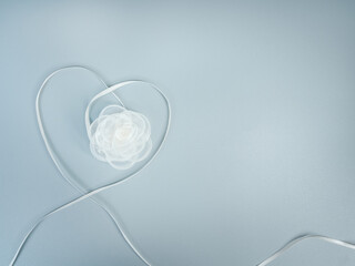 White satin ribbon forming a heart shape with delicate white rose on pastel blue background, symbolizing wedding, bridal theme, romance, love, anniversary, and elegant romantic celebration.