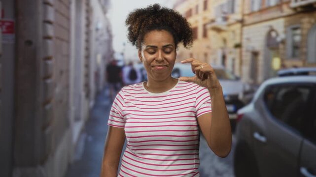 Woman wearing a pink striped shirt pinching thumb and index finger to show a small size while standing on a city street with parked cars; playful teasing.