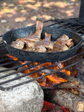 Pescado entero fresco fri&eacute;ndose en una sart&eacute;n de hierro fundido sobre una hoguera de le&ntilde;a, con aceite hirviendo y brasas brillantes, mostrando la cocina r&uacute;stica al aire libre
