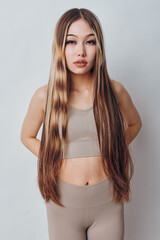Young woman with very long straight hair wearing beige sportswear standing against light background with serious expression and hands behind back in fashion portrait.