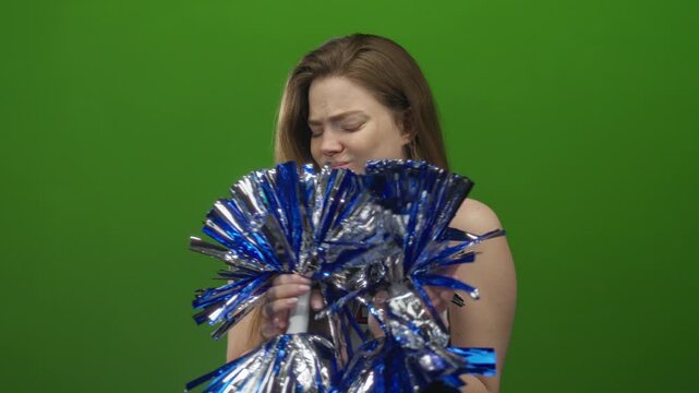 Cheerleader grips metallic blue pompons with both hands in green studio while grimacing at shiny streamers; disgust.