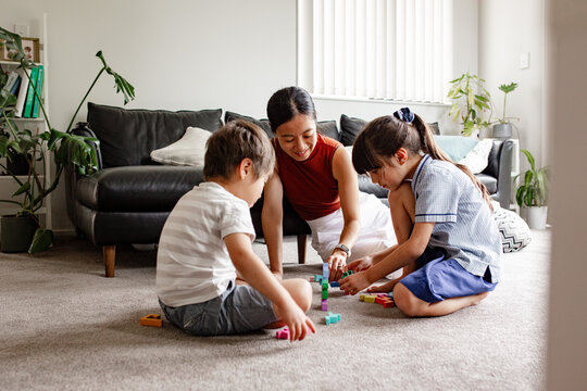 Two children seated on carpeted floor playing with blocks with mum nearby in living room