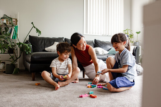 Two children seated on carpeted floor playing with blocks with mum nearby in living room