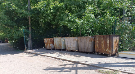 Row of heavily rusted, wheeled industrial dumpsters sits on a dusty patch of ground, backed by lush green trees and a wire fence under bright sunlight.