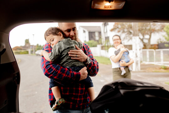 Father standing behind open car boot carrying sleeping toddler son