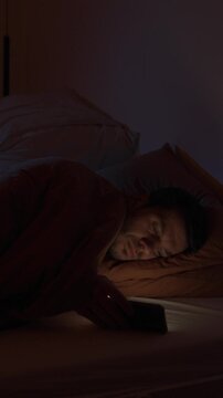 Man lying in bed holding smartphone and looking at screen in night. Concept of insomnia, sleep problems, night phone use and digital addiction