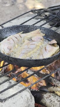 Pescado entero fresco fri&eacute;ndose en una sart&eacute;n de hierro fundido sobre una hoguera de le&ntilde;a, con aceite hirviendo y brasas brillantes, mostrando la cocina r&uacute;stica al aire libre