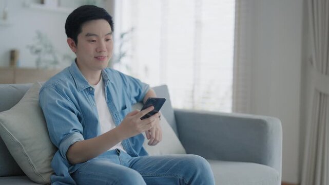 Smiling handsome young Asian man sitting on cozy couch, using funny mobile apps in living room. man at home, doing online shopping, messaging friends, posting on social media